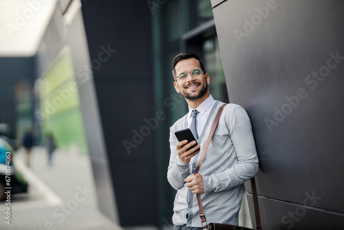 A cheerful young businessman elegantly dressed is standing at financial district and using his phone while smiling at the camera.