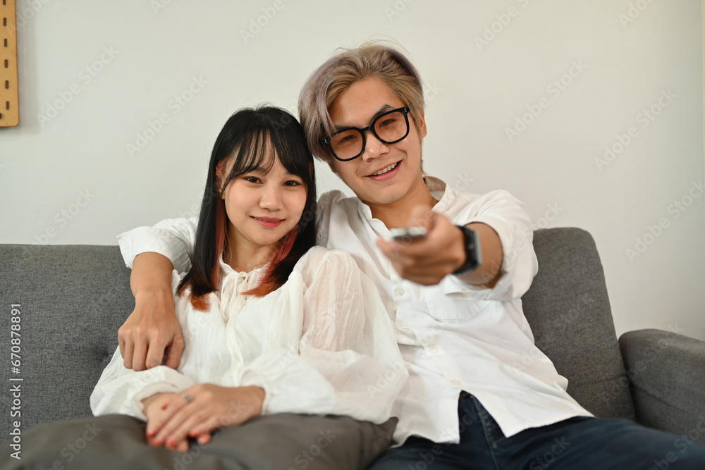 Happy young couple with remote control sitting on sofa at home watching television on weekend