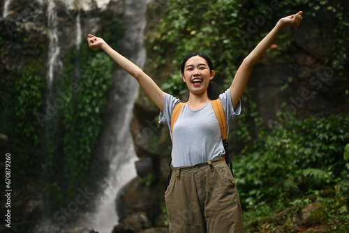 Cheerful female traveler with backpack raising arms enjoying the view of beautiful tropical waterfall