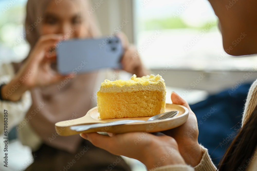 Cropped image of young woman holding plate of cake and her friend taking photo with smartphone