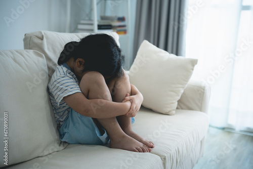 Asian kid girl upset lonely bullied little Sitting hugging your knees on the sofa couch in living room at home feels abandoned abused, sad alone, charity adoption concept. photo