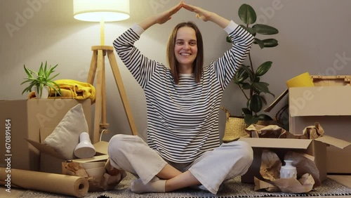 Young tenant. New house. Mortgage move. Adult smiling woman wearing striped shirt sitting on floor with her stuff and making roof with hands above her head photo