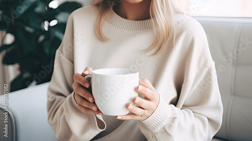 Blonde woman wearing light colored sweater sitting on comfortable sofa and holding blank white mug cup filled with coffee created with Generative AI Technology