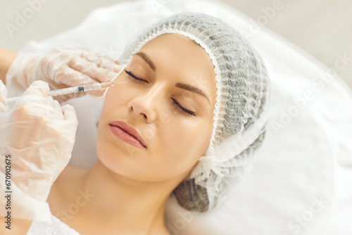 Young lady receives filler injection in lower eye lid area done by specialist at beauty clinic. Closeup woman's head in medical cap, hands in gloves holding syringe. Aesthetic facial treatment concept photo