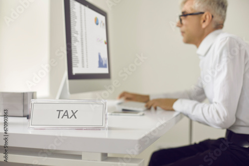 White sign with the word TAX on the table on the background of a serious busy senior man who works sitting at a laptop and checks the register of taxpayers. Business and finance concept.