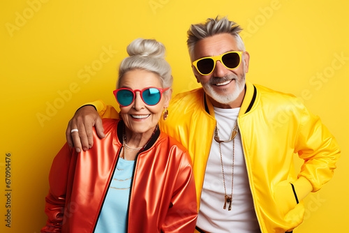 Stylish and fashionable elderly man and woman posing in bright jackets and sunglasses on a yellow background. Athleisure style