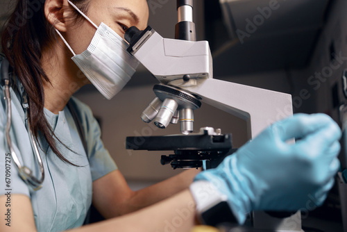 Asian doctor with medical mask and gloves looks into microscope at table in laboratory