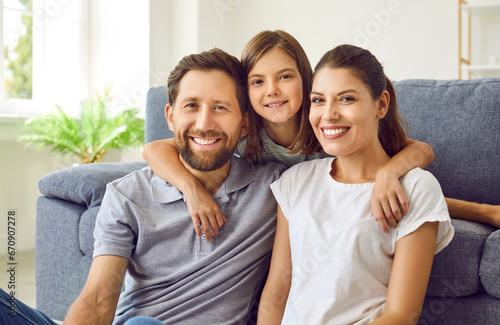 Close up portrait of cheerful smiling happy family sitting in the living room at home with their child girl and looking at camera. Cute daughter hugging her parents. Love and family leisure concept. © Studio Romantic