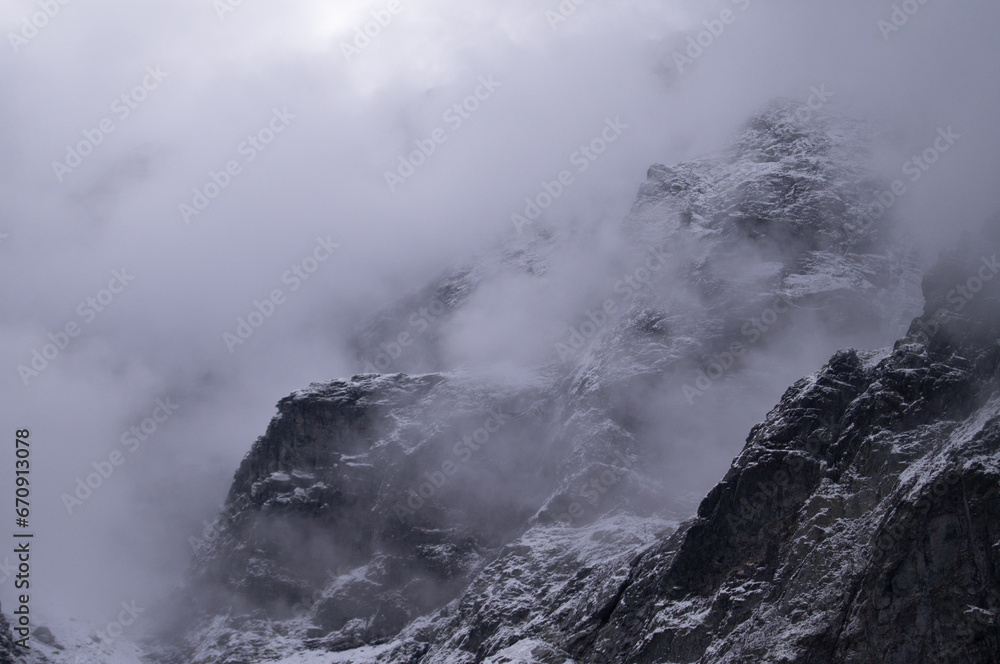 Captured in Rysy, this shot reveals the majesty of a snow-draped mountain peak, partially hidden by swirling mists. The rugged, icy surface and the ethereal fog present a stark, haunting beauty.