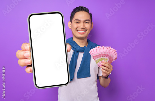 Cheerful young Asian man in white t shirt showing blank screen mobile phone and holding money banknotes isolated on purple background