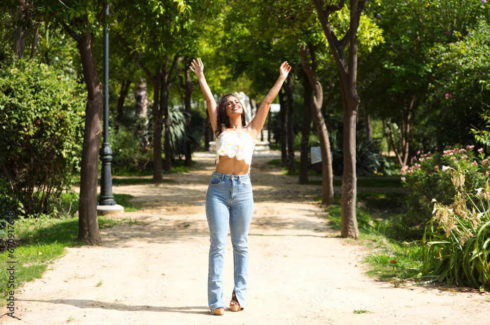 Obraz premium Beautiful Spanish brunette woman with long curly hair raises her arms to the blue sky. The woman is happy and thanks god and life for every day. Concept happiness and thankfulness.