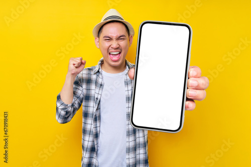 excited handsome Asian young man 20s wearing casual clothes hat Showing mobile cell phone with blank screen and making winner gesture isolated on yellow background. People lifestyle concept photo