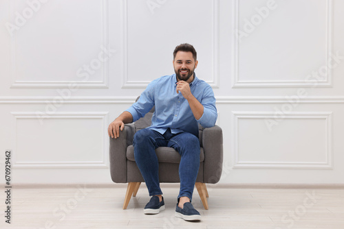 Handsome man sitting in armchair near white wall indoors