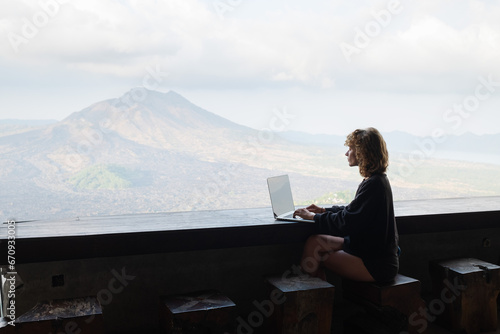 A young girl of European appearance is working on a laptop in a cafe with a gorgeous view of the mountains.
