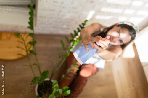 Young woman spraying water on potted plant indoors