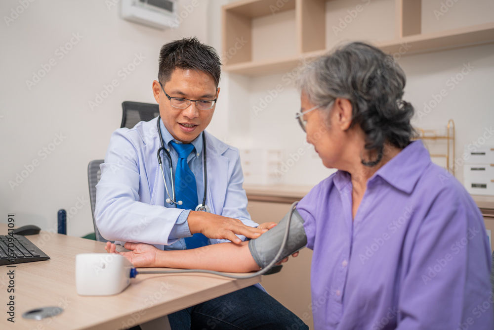 Fototapeta premium Asian caregiver doctor using digital pressure gauge measuring blood pressure of elderly woman patient. Young female nurse in uniform gives consultation during home visit. Medical and health concept.