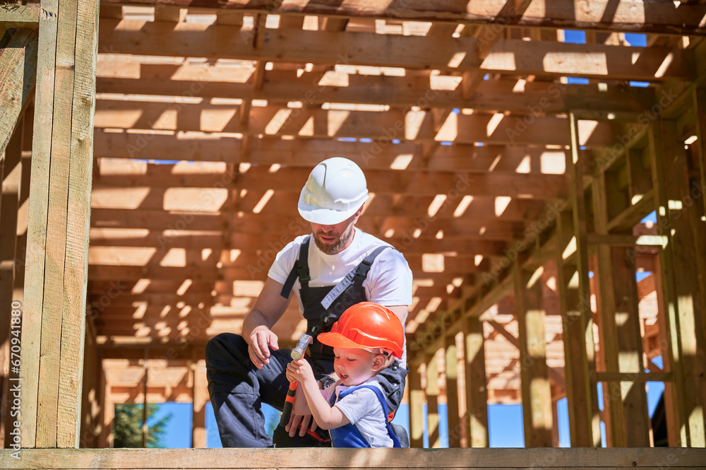 Father with toddler son building wooden frame house. Son attempting to hammer nail under guidance of his dad on construction site, wearing helmets and overalls on sunny day. Carpentry, family concept. Father with toddler son building wooden frame house. Son attempting to hammer nail under guidance of his dad on construction site, wearing helmets and overalls on sunny day. Carpentry, family concept.