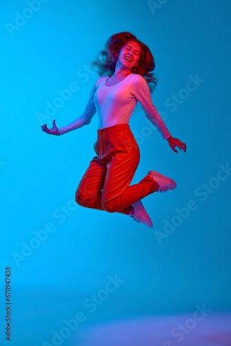 Full-length image of cheerful, positive, happy young girl jumping against blue studio background in neon light. Concept of youth, human emotions, facial expression, lifestyle © master1305