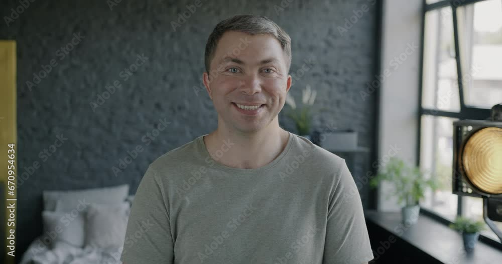 Slow motion potrait of attractive carefree man smiling looking at camera standing in modern bedroom with large window. People and emotion concept. Slow motion potrait of attractive carefree man smiling looking at camera standing in modern bedroom with large window. People and emotion concept.