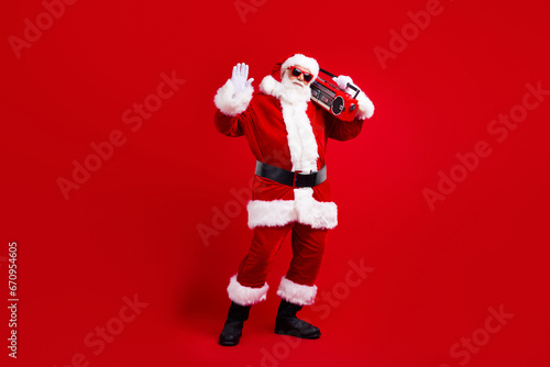Full size photo of cheerful grandfather wear santa costume in glasses hold boombox on shoulder say hello isolated on red color background © deagreez