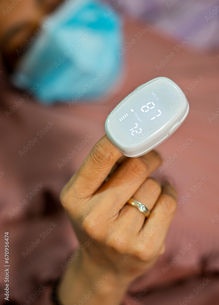 Fototapeta premium Pulse oximeter on a woman finger. A patient in a medical mask measures blood saturation with an electronic pulse oximeter on his finger. low saturation of the blood is covid 19 symptom.