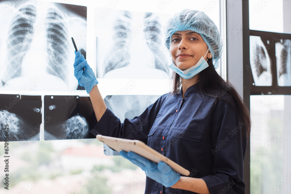 Indian female healthcare practitioner in protective uniform holding tablet with stylus pen while examining chest on x-ray indoors. Woman radiologist identifying mass in left lung using technologies.