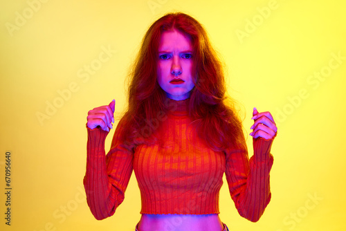Serious lookin girl standing with fists up and looking at camera against yellow studio background in neon light. Concept of youth, human emotions, facial expression, lifestyle