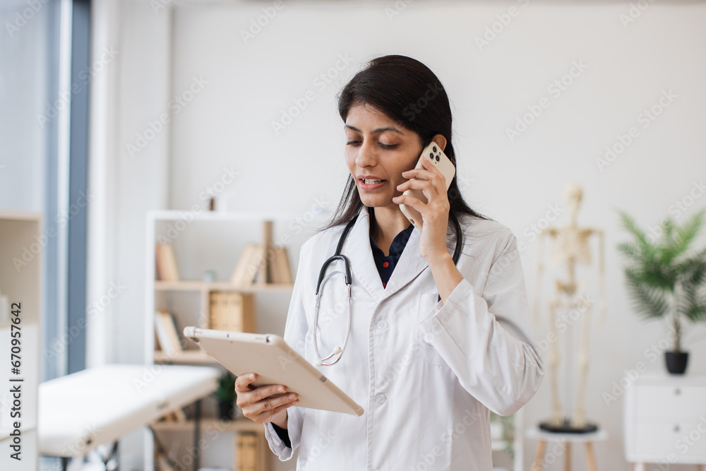 Front view of female physician standing and talking on mobile phone while looking on tablet screen. Competent hindu doctor using modern devices for work and consultation from hospital. Front view of female physician standing and talking on mobile phone while looking on tablet screen. Competent hindu doctor using modern devices for work and consultation from hospital.