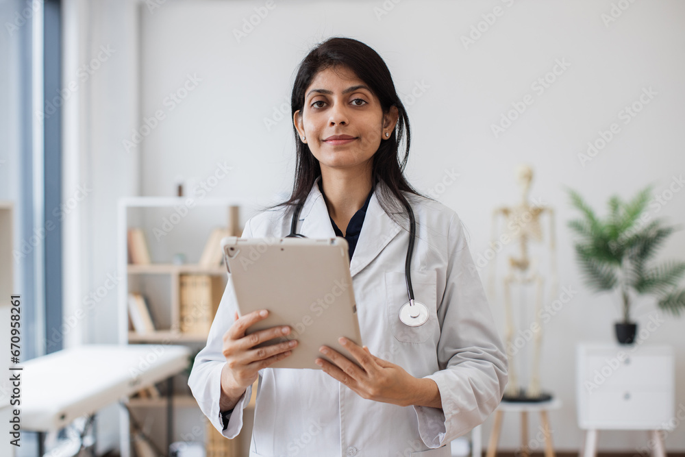 Portrait of friendly female physician in lab coat holding digital tablet and looking proudly at camera. Indian woman working in hospital using modern gadgets. Concept of perfect medical service. Portrait of friendly female physician in lab coat holding digital tablet and looking proudly at camera. Indian woman working in hospital using modern gadgets. Concept of perfect medical service.