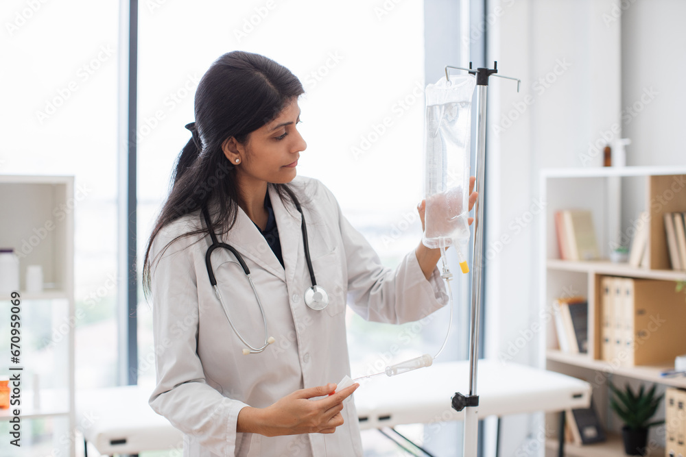 Adult female doctor in white lab coat standing near medical dropper and controlling liquid medication. Indian woman preparing infusion drip for intravenous treatment system.