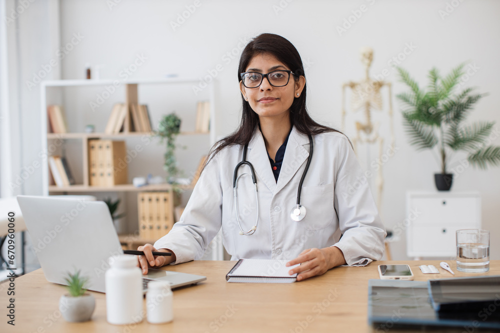 Female general practitioner in medical gown sitting and keeping hand on notebook while working with wireless laptop. Indian woman performing task professionally in field of medicine at office. Female general practitioner in medical gown sitting and keeping hand on notebook while working with wireless laptop. Indian woman performing task professionally in field of medicine at office.
