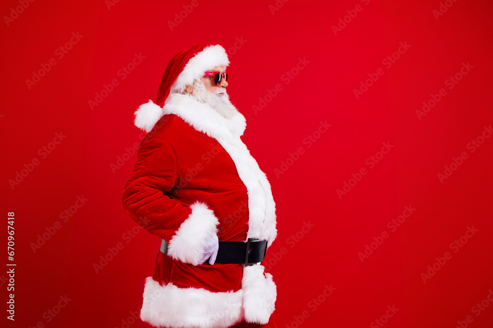 Side profile photo of confident grandfather wear santa costume look at empty space hands on waist isolated on red color background