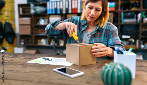 Blonde woman opening cardboard box with cutter in her local store. Female with freckles unpacking order received by courier with merchandise to sell on line. Parcel delivery and ecommerce concept.