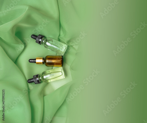 Three glass cosmetic bottles with oil and serum with a pipette on a green background of silk fabric with folds, a banner with an empty space for text, copy space, flat lay, skin care concept, spa
