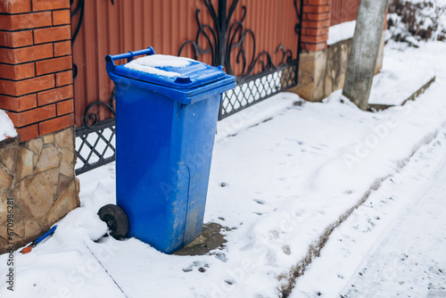 Close up view trash can covered with snow recycling containers with rubbish in front of gate private house. Winter garbage removal, household waste
