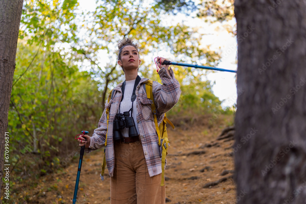 A young woman with curly hair in her 20s exploring the forest trail during autumn, wearing warm clothing and equipped with a yellow backpack, trekking poles, and binoculars. A young woman with curly hair in her 20s exploring the forest trail during autumn, wearing warm clothing and equipped with a yellow backpack, trekking poles, and binoculars.
