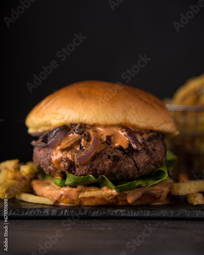 Juicy Homemade burger with lettuce onions, and chipotle sauce in a brioche bun. Dark and moody background. French Fries in background.