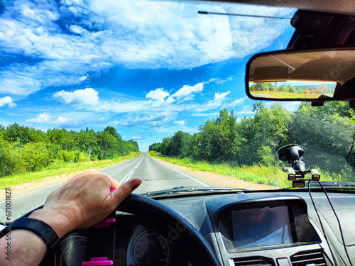 Car salon, steering wheel, hand of woman and view on nature landscape. View from seat of driver on Road, forest, blue sky, white clouds at sunny day. Concept of single trip of female traveller