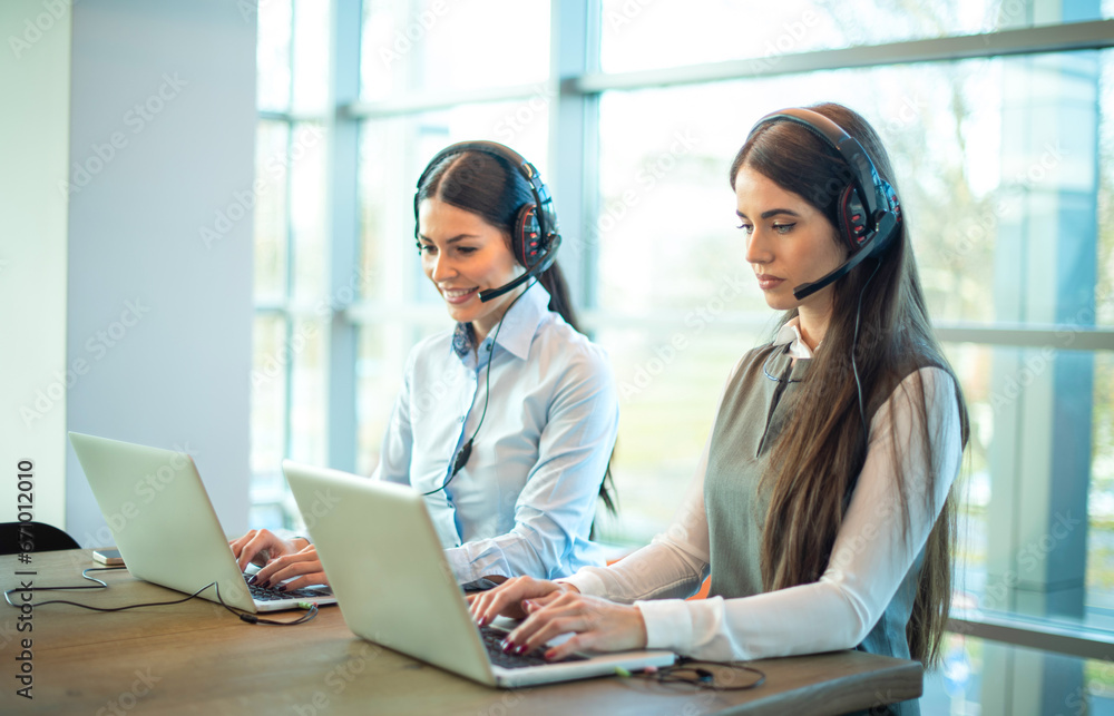 Two beautiful businesswomen at call center using laptops in corporate office. Attractive female employee workers wearing headset sitting on table using laptop, talking to customer at modern office. Two beautiful businesswomen at call center using laptops in corporate office. Attractive female employee workers wearing headset sitting on table using laptop, talking to customer at modern office.