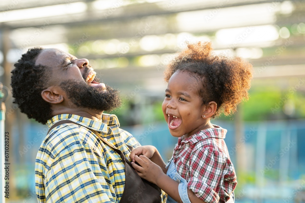 Happy african black parents dad father with daughter child on piggyback teasing fun in garden greenhouse. Black daughter kiss cheek dad and neck riding in vegetable greenhouse garden in light sunset - obrazy, fototapety, plakaty 