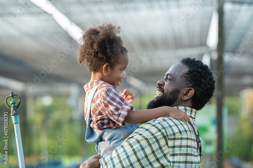 Happy african black parents dad father with daughter child on piggyback teasing fun in garden greenhouse. Black daughter kiss cheek dad and neck riding in vegetable greenhouse garden in light sunset