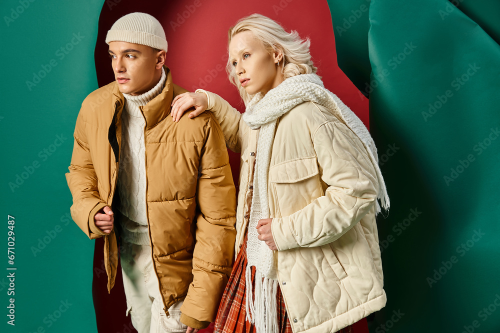 Fototapeta premium stylish man in beanie and puffer jacket posing with blonde woman on red with turquoise backdrop