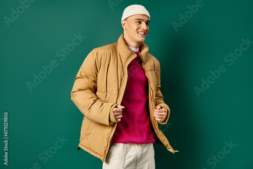 cheerful young man in beanie hat and stylish puffer jacket on turquoise background, winter trends photo