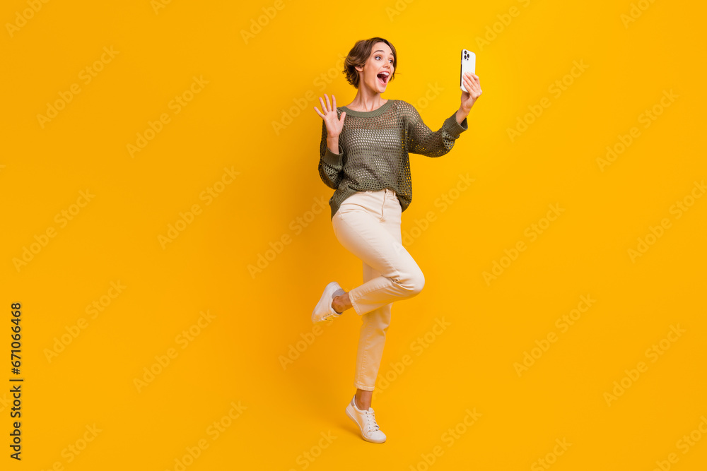 Full length photo of cheerful charming girl wear stylish sweater speaking online saying hello isolated on yellow color background