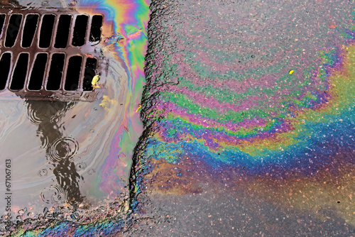 An oil slick against the backdrop of an asphalt road flows into a storm drain through a grate.
