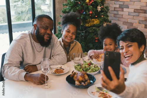 Cheerful african american family enjoying christmas and new years lunch while woman taking selfie on smartphone with members of family at home against decorated xmas tree