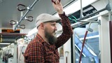 Tired millennial holds handrail in subway and looks into distance. Close-up of bearded millennial in cap goes at home after long day, carriage doors open at station. Concept millennial man use subway