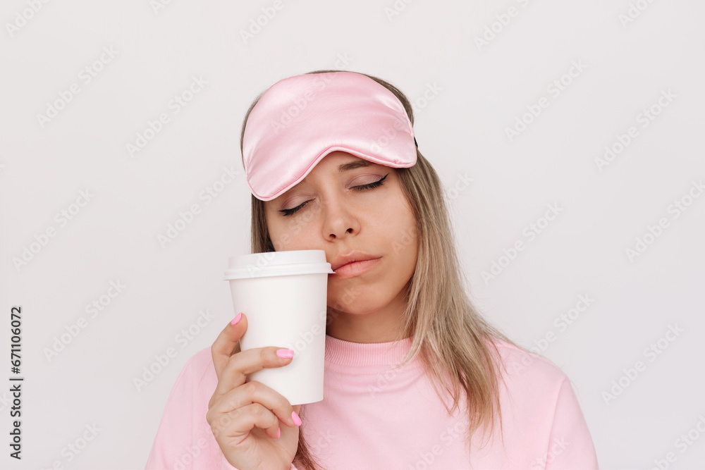Young sleepy blonde woman in a pink pajamas and sleep mask with her eyes closed holding white paper cup of coffee or tea in early morning on light background. Girl drinking coffee trying to wake up Young sleepy blonde woman in a pink pajamas and sleep mask with her eyes closed holding white paper cup of coffee or tea in early morning on light background. Girl drinking coffee trying to wake up