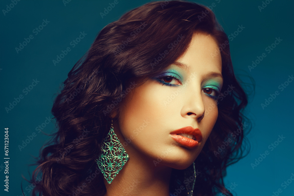Fototapeta premium Epoque of Disco concept. Close up portrait of fashion model with curly hair and arty make-up posing over blue background. Vintage turquoise earrings. Perfect skin. Retro style. Copy-space. Studio shot