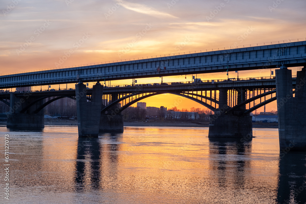 Beautiful view of the bridge over which cars drive at sunset. A river flows under the bridge, reflecting the sunset rays. Bridge in the city of Novosibirsk, Russia Beautiful view of the bridge over which cars drive at sunset. A river flows under the bridge, reflecting the sunset rays. Bridge in the city of Novosibirsk, Russia